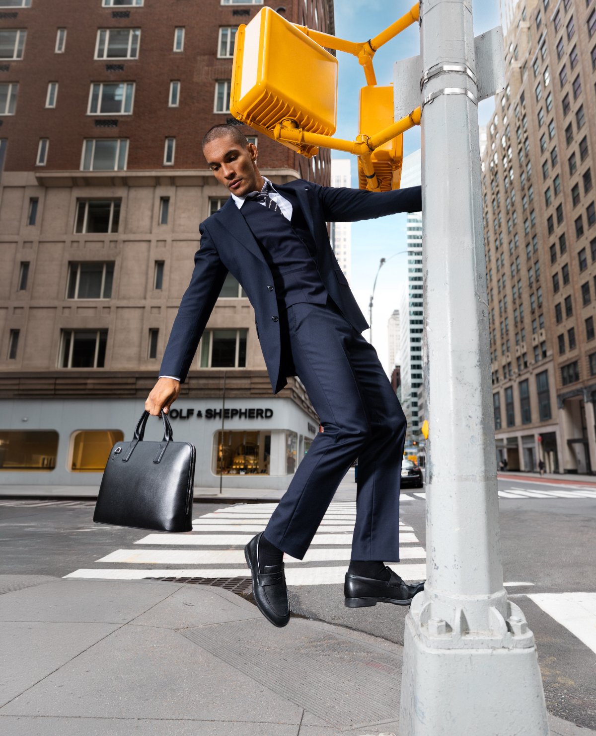 Man in a navy suit swinging off a crosswalk signal pole in black Capital Loafers