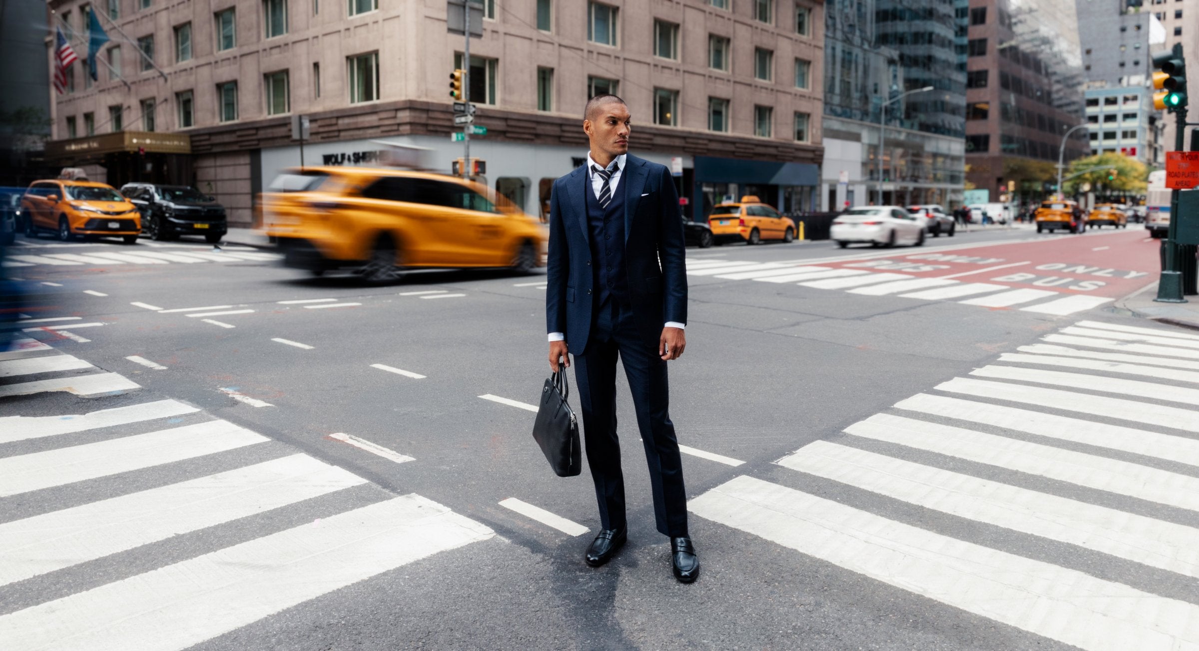 Man in a navy three-piece suit standing in a Manhattan crosswalk as a taxi passes