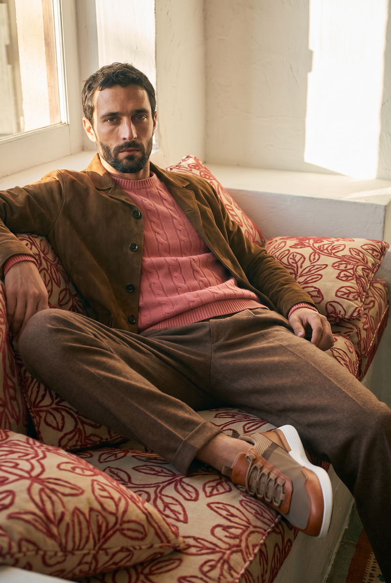 Model on a patterned sofa wearing Crossover Victory Trainers in Khaki / Honey