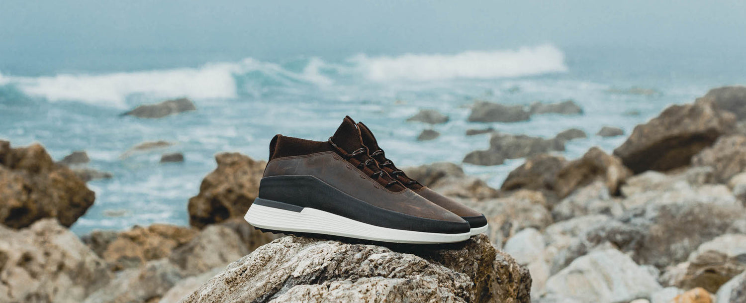 Brown boot with white sole resting on a rock in front of a wave crashing in a coastal scene. 