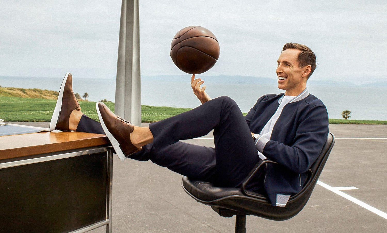 Man spinning basketball in index finger while leaning back in an office chair with his feet up on a desk. He is wearing leather dress shoes. 