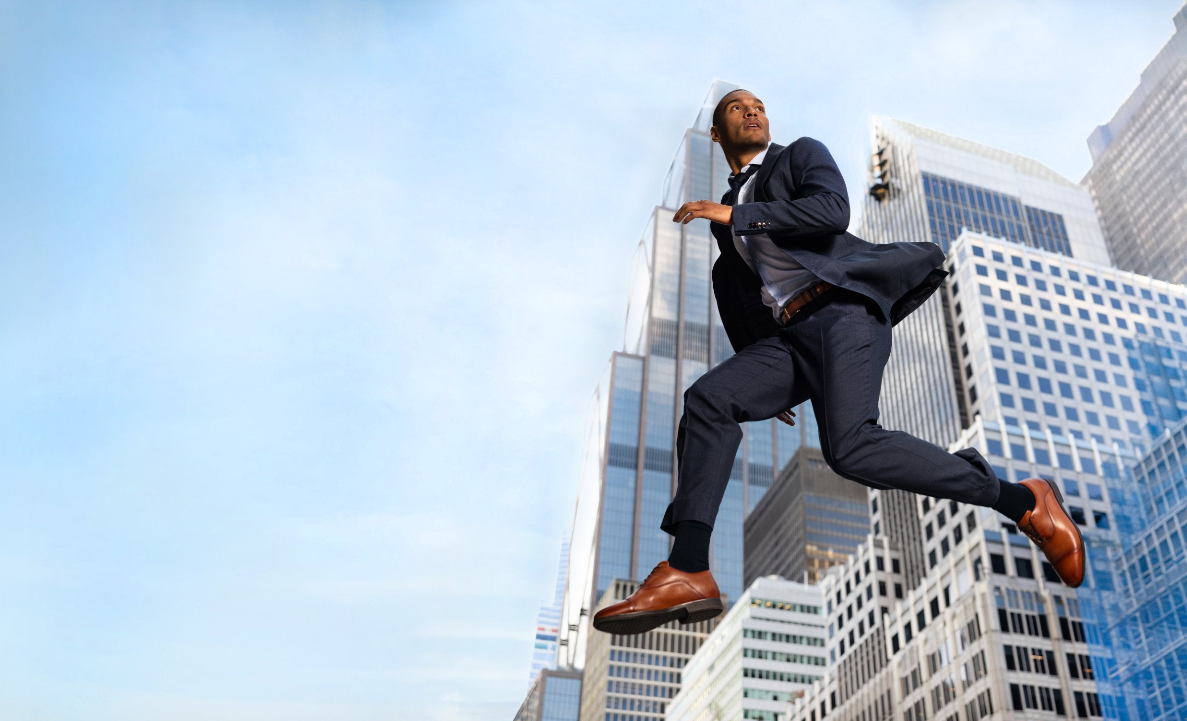Man in a navy suit leaping between Manhattan skyscrapers in whiskey Capital Leather Oxfords