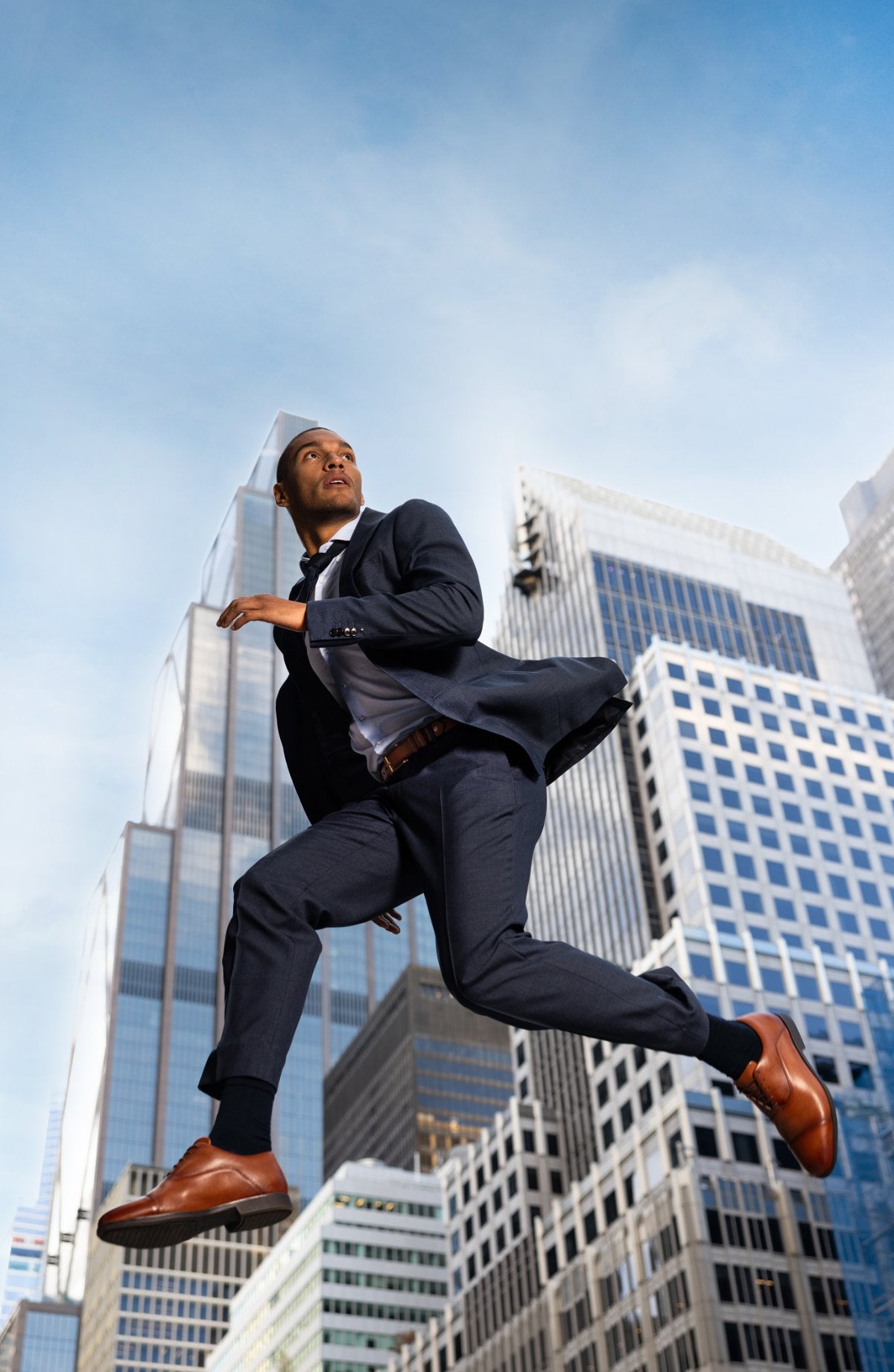 Man in a navy suit leaping between Manhattan skyscrapers in whiskey Capital Leather Oxfords