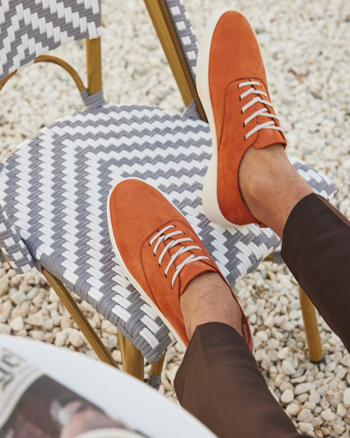 Orange, leather shoes resting on top of a blue and white woven chair on rocks at a cafe. 